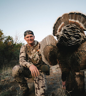 Janis Putelis kneeling beside a large wild turkey, wearing camouflage and a backward cap