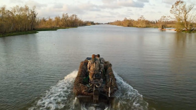 Two hunters on camouflaged blind boat traveling down a marshy bayou river