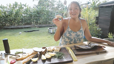 Woman holding a bite of toast, smiling at outdoor table with grilled fish and lemon wedges