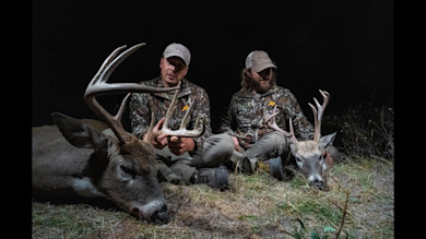 Two hunters at night seated with two bucks; left hunter holds large antlers