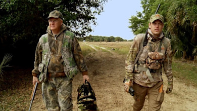 Two hunters in camouflage on a dirt road, one carrying a shotgun and duffel bag