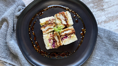 Chicken katsu sandwich cut into four squares with shredded slaw on a black plate ringed with sauce