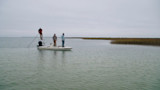 Three men fishing from a small skiff in shallow marsh under overcast sky