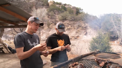Two men grilling meat on a riverside grate; one wears a MeatEater logo t-shirt