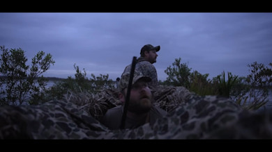 Two hunters in camouflaged blind at dusk, one standing looking away, one crouched with shotgun.