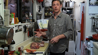 Steven Rinella holding a knife preparing a venison blade roast on a cutting board in a small kitchen