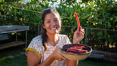 Woman smiling holding bowl of dried aku strips and one strip in hand outdoors