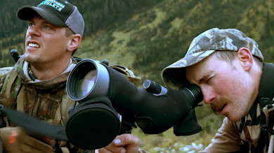 Two hunters in camouflage, one looking through a spotting scope in a mountain valley