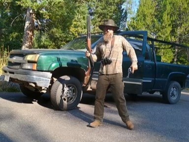 Man holding a shotgun upright in front of a green pickup truck parked on a road