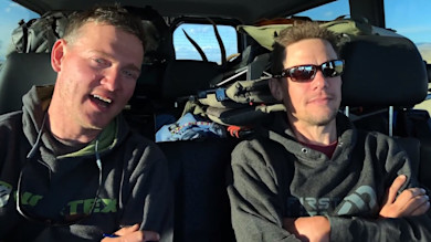Two men in vehicle backseat with folded hunting gear; man on right wearing sunglasses