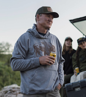 Man in cap and grey hoodie with bison graphic holding yellow can beside two people and YETI cooler