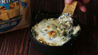 Cheesy stinging nettle and artichoke dip in a black skillet, cracker scooping; chip bag reads SIMPLE INGREDIENTS