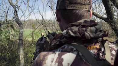 Hunter from behind in camouflage jacket holding a turkey box call; 'IRSTL' text on pack