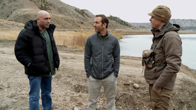 Three men standing on a rocky riverbank with hills; one wears camouflage chest pack