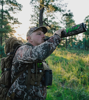 Hunter blowing into camo Phelps call in pine forest at sunset