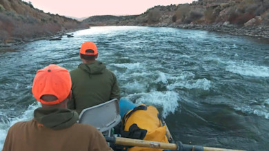 Two hunters in orange caps rafting through rapids on a canyon river