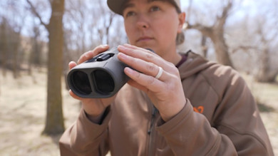 Person outdoors holding gray binoculars toward camera, trees in background