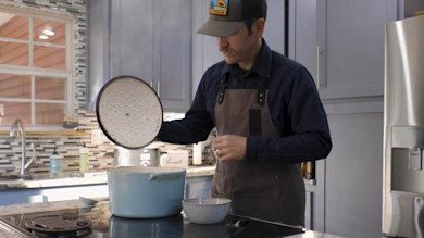 Man in cap and apron lifts lid from blue pot on stove; bowl beside; canister labeled 'bread'
