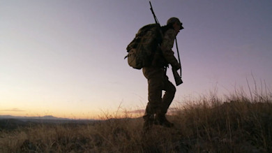 Silhouetted hunter walking up grassy hill at sunrise carrying rifle and backpack