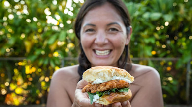 Fried fish slider held to camera with greens and sauce, woman smiling outdoors
