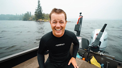 Man smiling on boat wearing black wetsuit labeled 'AKONA ADVENTURE GEAR' with outboard motor on lake