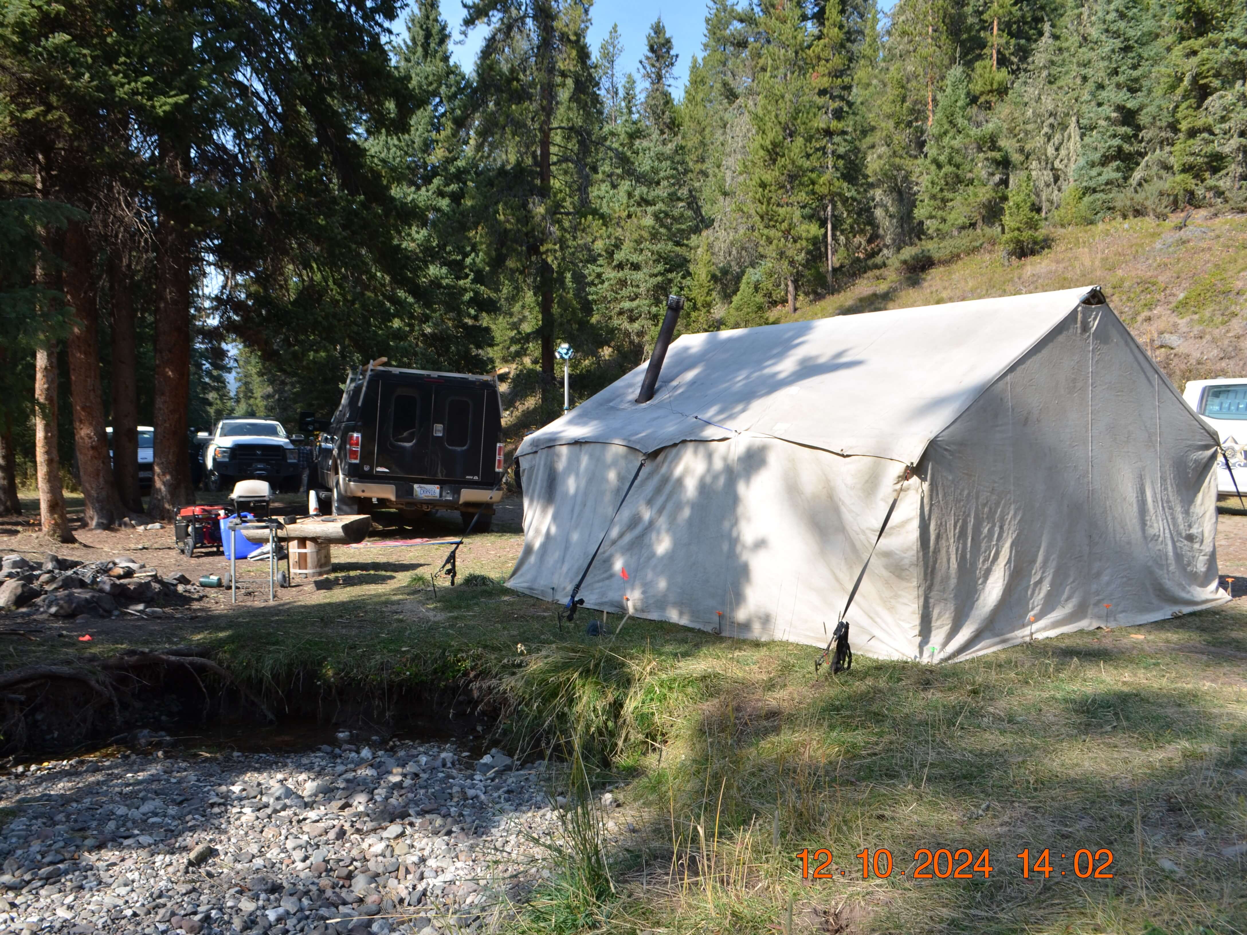 Canvas tent and trucks at forest campsite; timestamp 12.10.2024 14:02