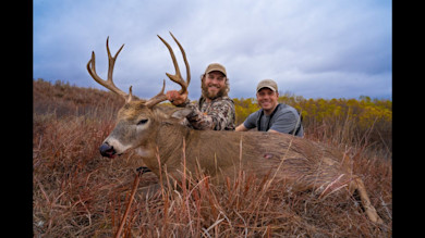 Downed buck with large antlers; two hunters kneel behind it in brown grass