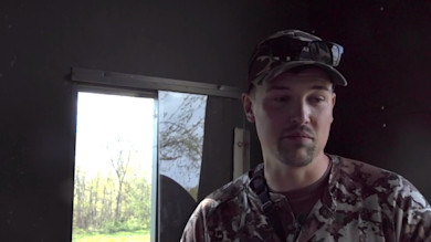 Hunter in camouflage inside a hunting blind, looking toward a bright window