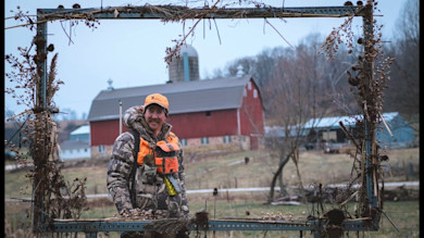 Hunter in camouflage jacket and orange vest framed by rusted metal frame with red barn behind