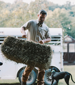 Man lifting a hay bale beside a white trailer with a black dog nearby