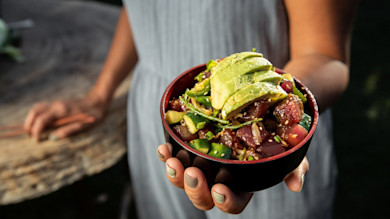 Poke bowl topped with sliced avocado, diced tuna, cucumber and sesame, held by person