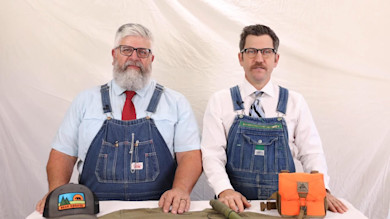 Two men in denim overalls and ties seated at a table with cap labeled "Bear Grease" and orange pouch