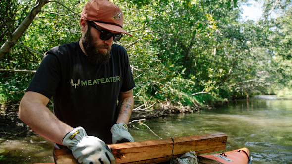 Bearded man wearing black MEATEATER tee handling a wooden plank on an orange kayak