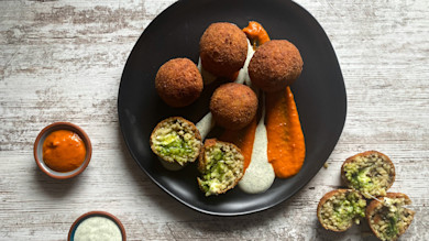 Five fried arancini on black plate with orange and white sauce streaks; two halved show green filling