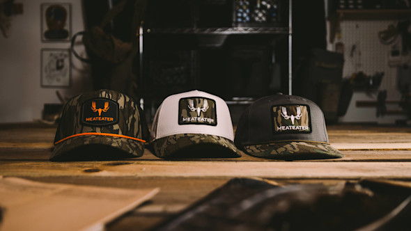 Three MeatEater trucker hats—left camo with orange trim, center white, right gray—with camo patches reading MEATEATER