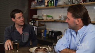 Steven Rinella and another man seated at table with plate of sausages, kitchen shelves, 'MEAT EATER' text