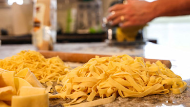 Fresh homemade pasta piled on floured countertop, rolling pin behind, blurred hands in background