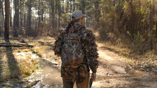 Hunter on forest trail wearing leafy camo vest, First Lite pack, turkey tail feathers over shoulder