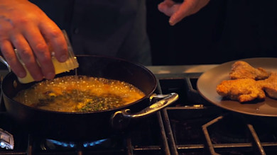 Bubbling piccata sauce in skillet as hand pours lemon; breaded turkey cutlets on plate
