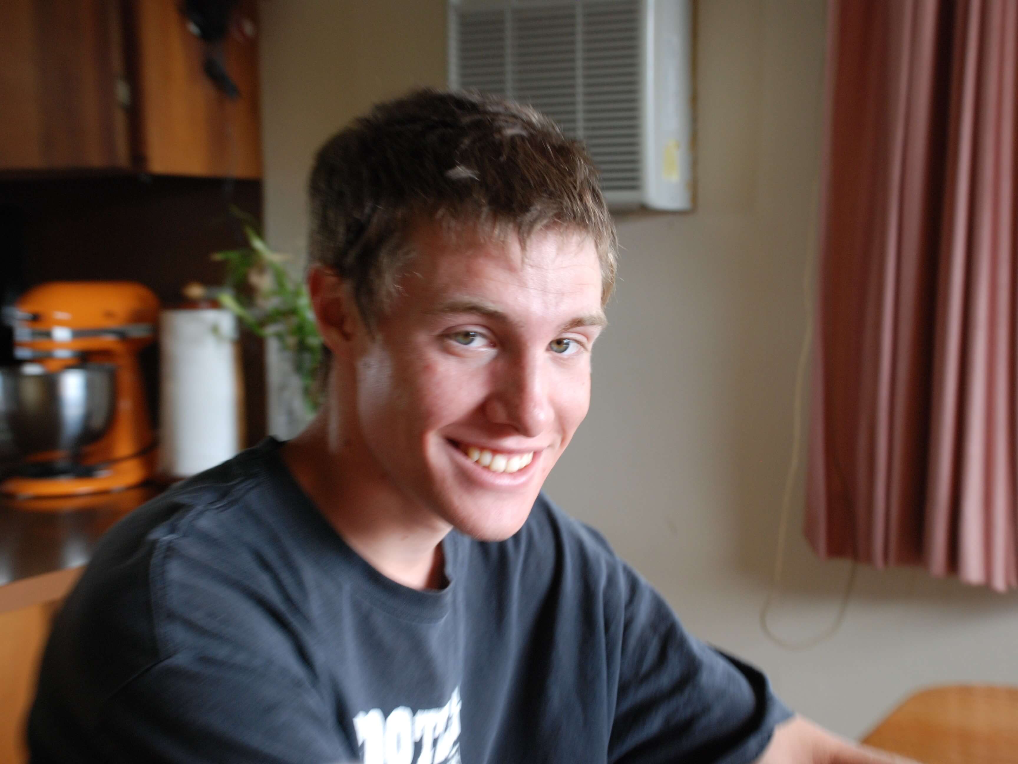 Young man smiling at kitchen table, orange stand mixer in background