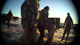 Three hunters carry two coyotes by tails beside a pickup in a snowy parking lot at sunset