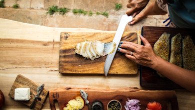 Thin sliced cold-smoked fish on wooden board; hands slicing seasoned fillets with capers, red onion, tomato, butter.