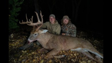 Two hunters kneel behind a large buck with massive antlers in a dark forest at night