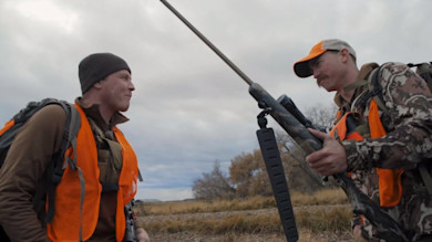 Two hunters on a riverbank, one holding a rifle upright between them, both wearing orange vests