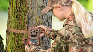 Young woman in camouflage attaching a brown trail camera to a tree with a strap