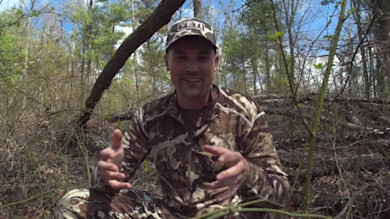 Hunter in camouflage and YETI cap gesturing while seated in leafless spring woods