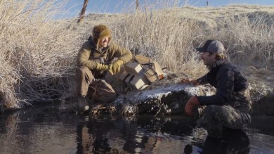 Two hunters kneeling by a frosty stream with gear, demonstrating a pocket-set trap