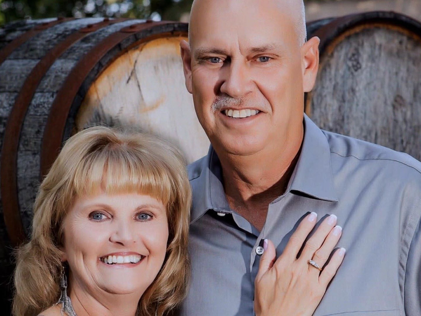 Smiling couple posed before wooden barrels; woman's hand with ring on man's chest