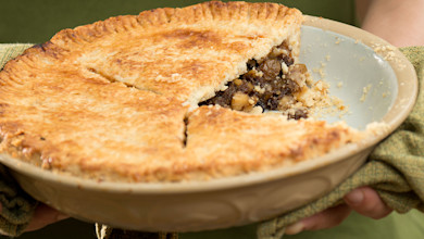 Golden-crusted wild-game mincemeat pie, one slice removed revealing dark meaty filling, hands holding pie dish