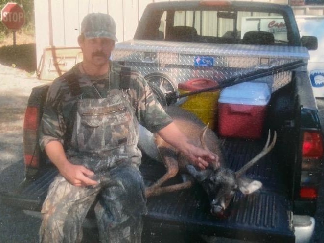 Hunter in camouflage overalls sitting on pickup tailgate beside a harvested buck with antlers, STOP sign behind